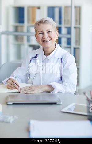 Ritratto di medico maturo femmina sorridente alla macchina fotografica mentre siede al posto di lavoro nell'interno della clinica Foto Stock