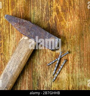 Vecchio martello arrugginito e tre chiodi in acciaio giacciono su una tavola di legno graffiata. Spazio di copia per il testo. Messa a fuoco selettiva. Concetto di lavoro domestico. Formato quadrato. In alto vi Foto Stock