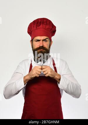 Cuoco con faccia rigorosa in uniforme di borgogna mostra posate. Concetto di ristorazione al ristorante. Lo chef attraversa le stoviglie. L'uomo con barba tiene forchetta e cucchiaio su sfondo bianco. Foto Stock