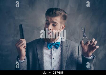 Ritratto di giovane uomo bearded di stile in un vestito con fiocco-cravatta si alza su uno sfondo grigio e sceglie tra forbici e un pettine. Lui risolve andare a. Foto Stock