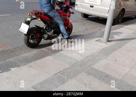 Motocicletta e conducente sulla strada. Il conducente sta aspettando con il piede sul marciapiede. La parte inferiore è fotografata. Foto Stock