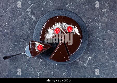 Torta al cioccolato nella foresta nera con ripieno di ciliegie con smalto al cioccolato fondente decorato con ciliegie in scatola e cioccolato bianco, una fetta posizionata Foto Stock