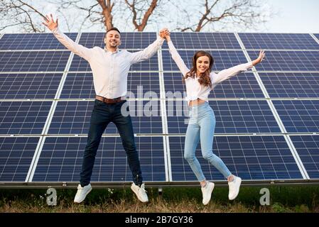 Allegro giovane coppia che salta insieme tenendo le mani sullo sfondo dei pannelli solari sotto un cielo blu. Giovani che indossano jeans e camicie bianche. Immagine del concetto di energia solare Foto Stock