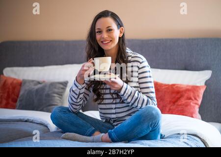 Giovane donna felice seduto a letto in casa sua e bere il caffè. Foto Stock