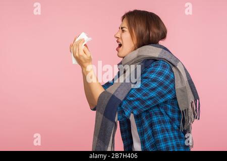 Influenza stagionale. Vista laterale della ragazza malata in camicia a scacchi avvolta in sciarpa che soffia naso runny, starnuti in tovkin, sofferenza infezione virale sintomi. i Foto Stock