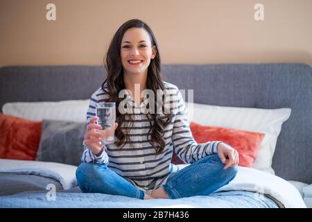 Giovane donna felice seduto a letto in casa sua, guardando la macchina fotografica e tenendo un bicchiere d'acqua. Foto Stock