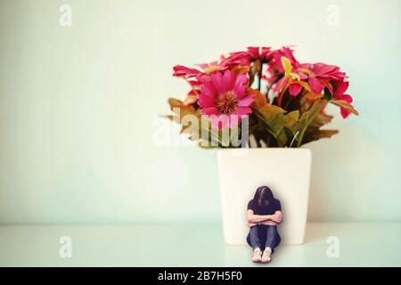 Triste donna depressa seduta con la schiena appoggiata contro bianco vaso con falso fiore rosso. Concetto minimo di tristezza, solitudine. Foto Stock