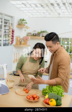 Bella coppia Giovane è utilizzando un telefono cellulare e sorridente durante la cottura in cucina a casa Foto Stock