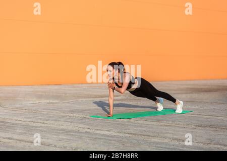 Atletica si adatta bella ragazza in abbigliamento sportivo, pantaloni neri e top, pratica yoga, in piedi in Phalakasana più alta posizione asse con una mano, muscolo di allenamento Foto Stock