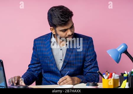 Ritratto di stanco pensivo giovane imprenditore indiano seduto sul posto di lavoro, tenendo la penna, pensando a problemi finanziari, indossando tuta, guardando però Foto Stock