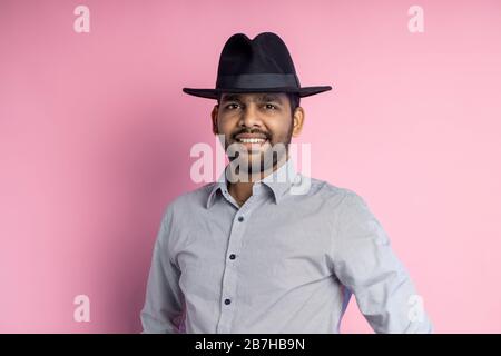 Primo piano di felice giovane indiano maschio con stoppia, indossare camicia casual, cappello nero, guardando con il sorriso e puntando a lato con forefinge Foto Stock