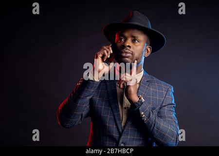 Scatto orizzontale di serio preoccupato giovane africano elegante vestito, cappello, parlando sul telefono cellulare, in piedi in buio su sfondo nero . Suc Foto Stock