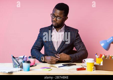 Inquadratura orizzontale di bello fiducioso pragmatico afro american businessman seduti al posto di lavoro in ufficio, pulsante di fissaggio sulla tuta, indossando occhiali Foto Stock