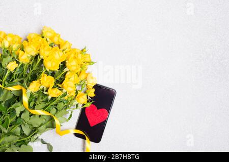 Fiori di rose gialle e telefono con cuore rosso su tavolo bianco sfondo vista dall'alto mockup spazio libero Foto Stock
