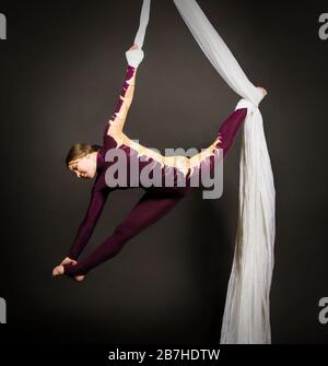 Donna sportiva in una tuta di Borgogna esegue ginnastica e circo esercizi su seta bianca. Riprese in studio su sfondo scuro, immagini isolate. Foto Stock