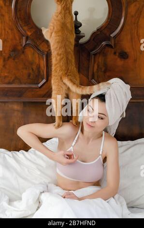 giovane donna con un gatto rosso sulla spalla, un asciugamano sulla testa si siede in un vecchio letto, sorridendo. Foto Stock