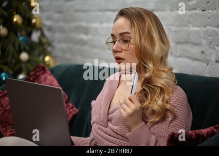 Lady blogger in cardigan rosa, pantaloni, occhiali. Seduto sul divano verde con cuscini colorati, mostrando carta di plastica, guardando il suo computer portatile. Primo piano Foto Stock