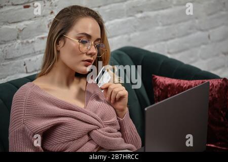 Lady blogger in cardigan rosa e occhiali. Si siede su un divano verde con cuscini colorati, ha una scheda di plastica e guarda il suo computer portatile. Primo piano Foto Stock