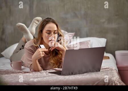 Blonde ragazza blogger in occhiali, abbigliamento casual. Appoggiandosi al mento, tenendo la carta di plastica, appoggiandosi sul letto con il laptop, il libro e la tazza. Primo piano, spazio di copia Foto Stock