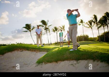 Uomo e due donne guardare un golfista maschio prendere un colpo su una trappola di sabbia. Foto Stock