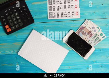 Calendario, notebook pulito, smartphone rotto e denaro russo sul tavolo blu. Le spese impreviste hanno interrotto i piani Foto Stock