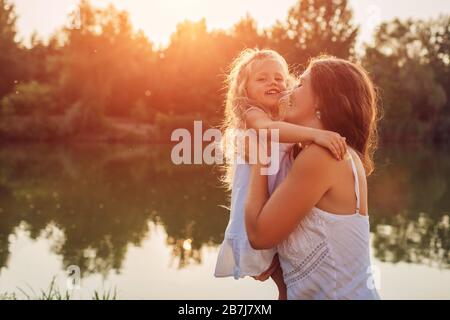 Giorno della madre. Donna che gioca divertendosi con la figlia sul fiume d'estate al tramonto. Donna che tiene il capretto e che ride. Famiglia Foto Stock