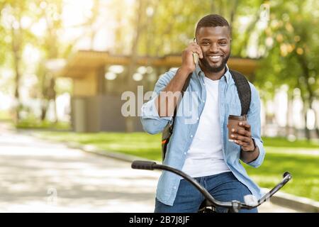 Un uomo nero sorridente in bicicletta che tiene il caffè e parla sullo smartphone Foto Stock