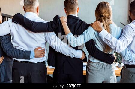 I colleghi che si levano in piedi indietro alla macchina fotografica si abbracciano durante la riunione in ufficio Foto Stock