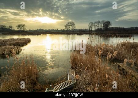 Un ponte tra canne sulla riva di un lago ghiacciato, il sole tramonta dietro le nuvole Foto Stock