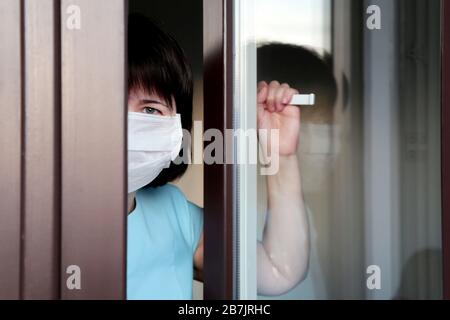 Quarantena del coronavirus durante l'epidemia di COVID-19. Donna triste in una maschera medica guarda fuori dalla finestra Foto Stock