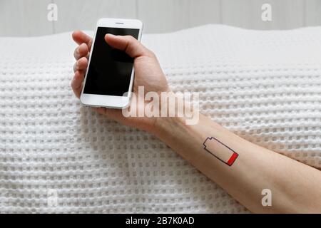 Simbolo di batteria scarica tracciato sul polso umano. Spossatezza uomo sdraiato sul letto, tenendo il cellulare in mano, vista dall'alto. Dipendenza da smartphone gadget, ch Foto Stock