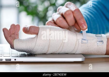 Primo piano delle braccia dell'uomo che avvolgono il suo polso doloroso con bendaggio ortopedico elastico di supporto flessibile causato da un lavoro prolungato sul laptop. Tunnel carpale Foto Stock