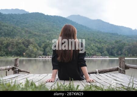 Immagine di primo piano di una donna asiatica seduta da sola su un vecchio molo di legno vicino al fiume con cielo e sfondo di montagna Foto Stock