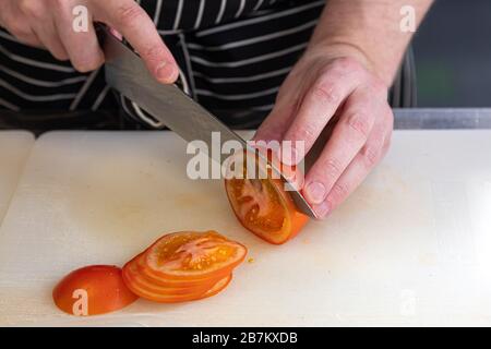 primo piano chef maschile in un grembiule nero con un coltello affilato taglia un pomodoro rosso in fette sottili su un pannello di plastica Foto Stock