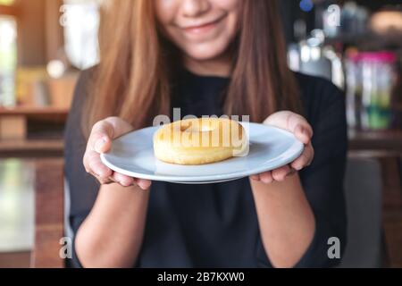 Una bella donna asiatica che tiene e mostra un piatto di ciambella con sentirsi felice e buon stile di vita nel moderno caffè Foto Stock