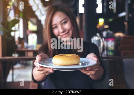Una bella donna asiatica che tiene e mostra un piatto di ciambella con sentirsi felice e buon stile di vita nel moderno caffè Foto Stock