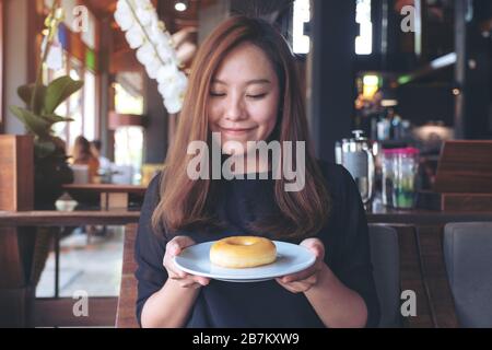 Una bella donna asiatica che tiene e mostra un piatto di ciambella con sentirsi felice e buon stile di vita nel moderno caffè Foto Stock