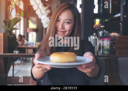 Una bella donna asiatica che tiene e mostra un piatto di ciambella con sentirsi felice e buon stile di vita nel moderno caffè Foto Stock