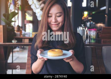 Una bella donna asiatica che tiene e mostra un piatto di ciambella con sentirsi felice e buon stile di vita nel moderno caffè Foto Stock