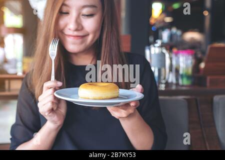 Una bella donna asiatica che tiene un pezzo di ciambella e forchetta con sentirsi felice in un bar Foto Stock