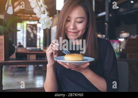 Una bella donna asiatica che tiene un pezzo di ciambella e forchetta con sentirsi felice in un bar Foto Stock