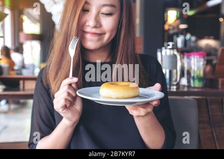 Una bella donna asiatica che tiene un pezzo di ciambella e forchetta con sentirsi felice in un bar Foto Stock