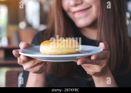Una bella donna asiatica che tiene e mostra un piatto di ciambella con sentirsi felice e buon stile di vita nel moderno caffè Foto Stock