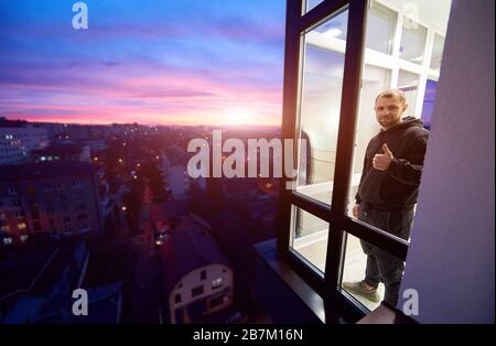 L'uomo è in piedi su un balcone con finestre panoramiche e mostrando un pollice su come segno con un tramonto magico fuori della finestra. Paesaggio urbano dall'alto e cielo pieno di fuoco multicolore con sole che tramonta Foto Stock