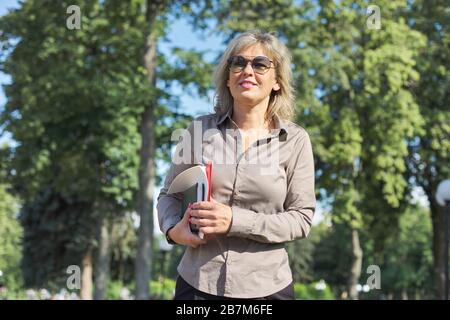 Donna matura e sicura per il business che cammina con documenti all'aperto. Bella donna bionda in occhiali, camicia grigia, sfondo Sky Park Foto Stock