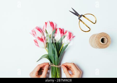 Le mani della donna che legano la corda di iuta si inchino su un semplice bouquet di fiori freschi di tulipani su sfondo bianco, disposizione piatta. Foto Stock