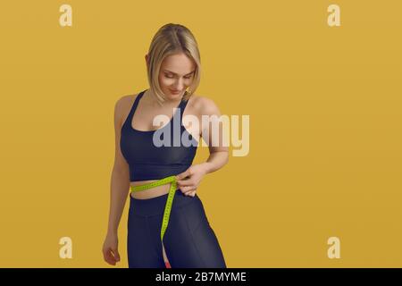 Giovane donna sottile e sportiva che misura la vita con un metro a nastro, mentre guarda verso il basso con un sorriso soddisfatto in una perdita di peso, dieta sana e esercizio fisico c Foto Stock
