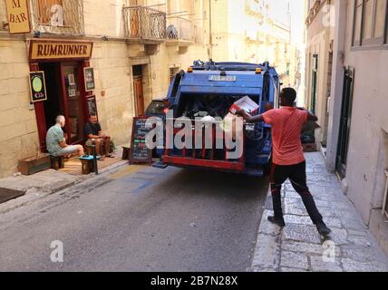 Valletta. Malta. Camion per la raccolta di rifiuti nella strada della città vecchia. L'uomo che raccoglie i sacchetti dei rifiuti è immigrato africano. Foto Stock