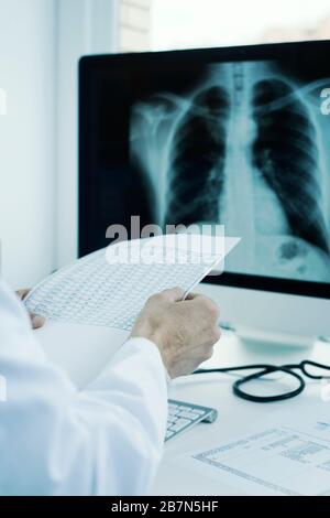 primo piano di un giovane dottore, con un cappotto bianco, seduto alla sua scrivania per controllare i risultati di una prova e una radiografia toracica nel suo computer Foto Stock