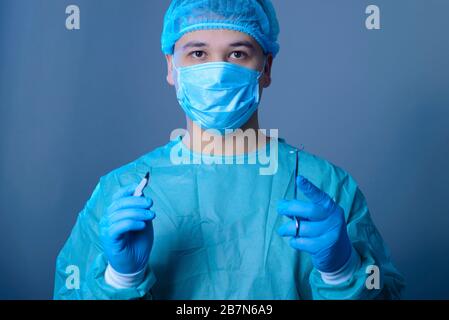 primo piano ritratto di un medico caucasico chirurgo, in una tuta sterile, maschera, tenendo un bisturi e supporto dell'ago con un ago, preparandosi ad operare. Acceso Foto Stock